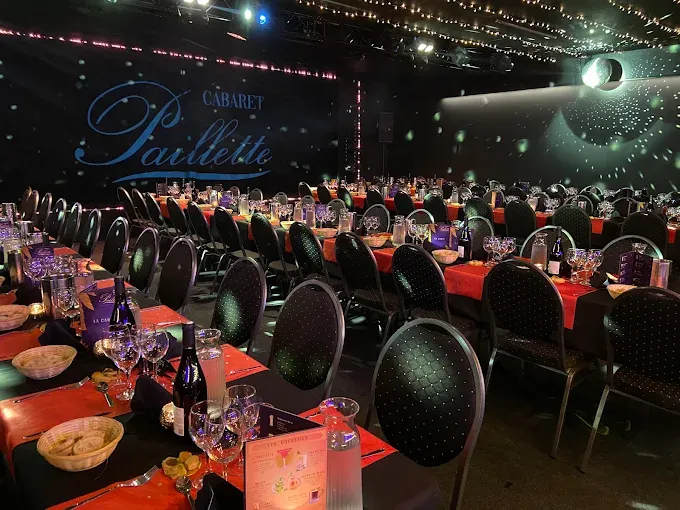 Salle du Cabaret Paillette avec les tables dressées, serviettes rouges, verres et chaises face à la scène, sous les guirlandes lumineuses.